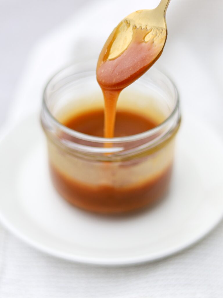 Gold spoon with homemade caramel sauce dripping from it into a clear glass jar.